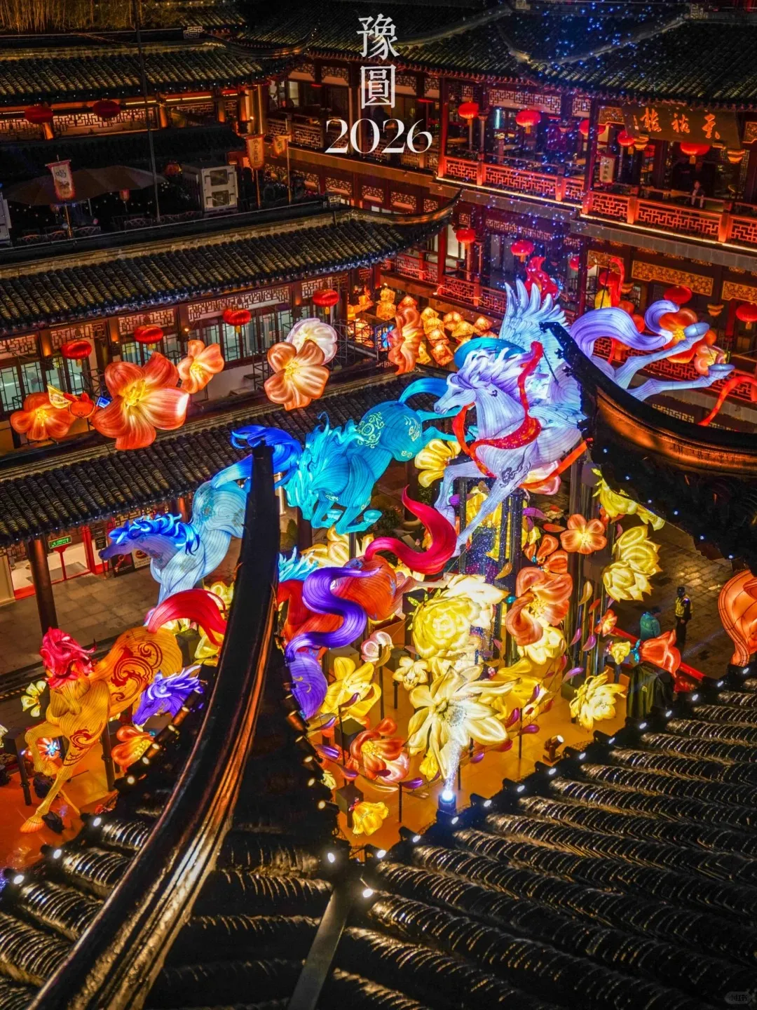 Overhead view of Yuyuan Garden in Shanghai at night, illuminated by hundreds of vibrant lanterns. Large, colorful lanterns shaped like mythical horses in blue, white, purple, and red ascend from the lower left. Surrounding them are many glowing flower-shaped lanterns in orange, yellow, and red. Traditional Chinese buildings with dark tiled roofs and red lanterns are visible in the background, with the Chinese characters '豫園' and '2026' displayed in white at the top.