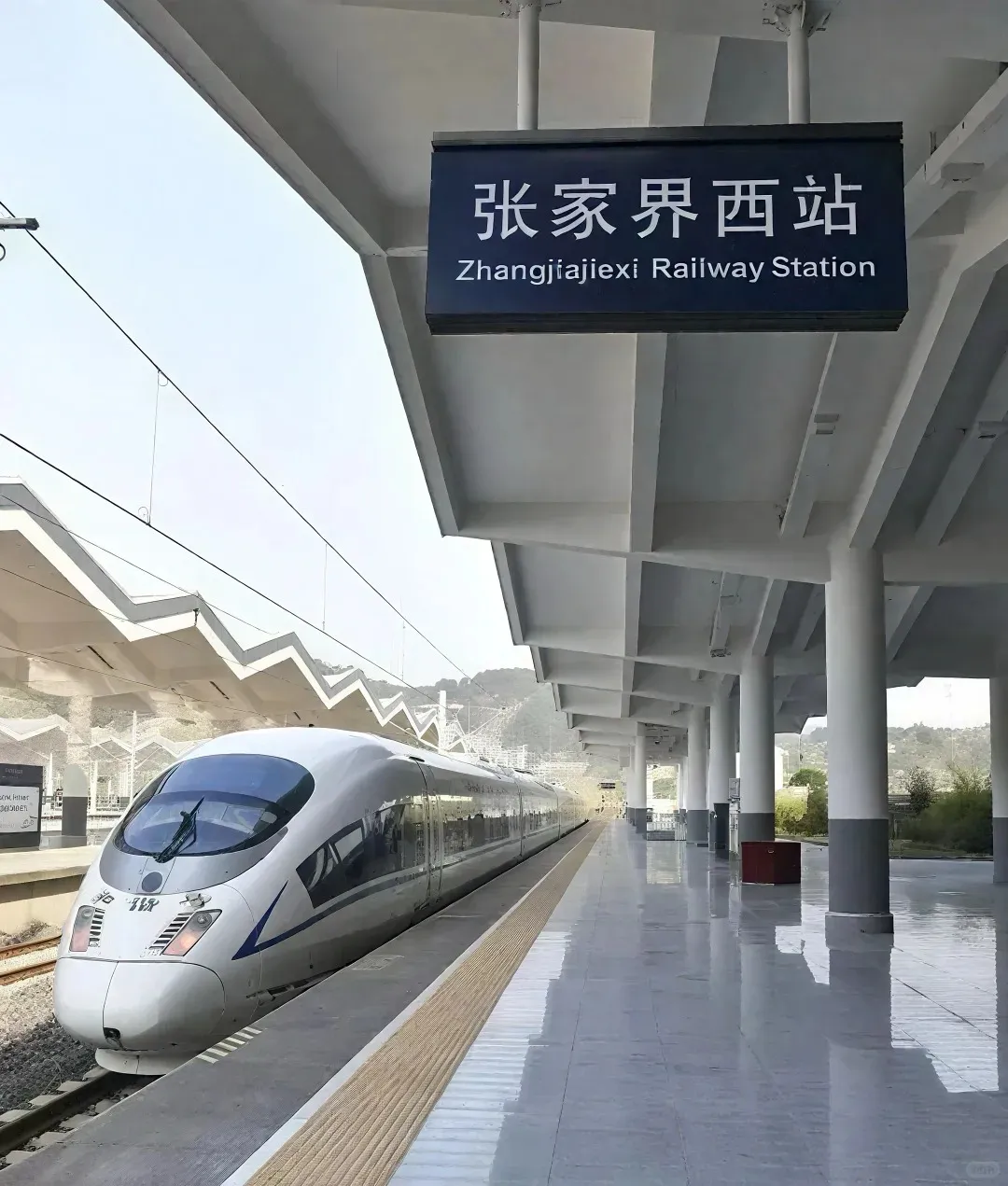A sleek white high-speed train is parked at the platform of Zhangjiajiexi Railway Station, indicated by an overhead sign in Chinese and English. The modern station has grey pillars and a patterned canopy, with distant green mountains under a clear sky.