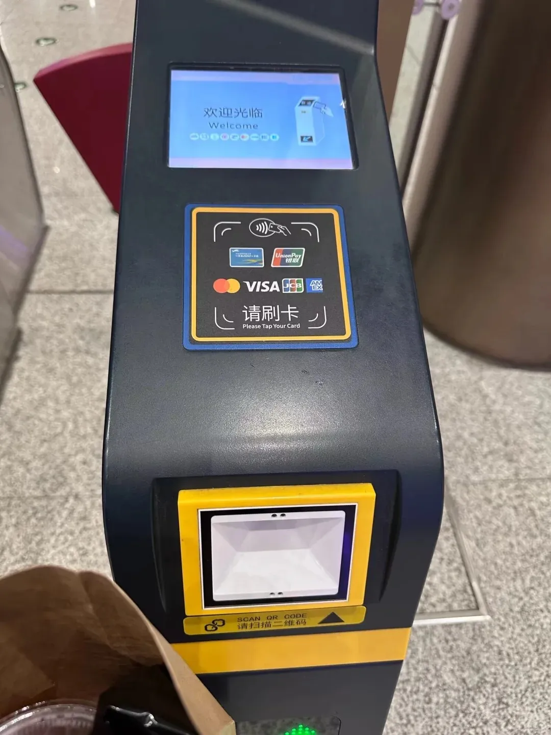 A dark grey and yellow payment terminal at a subway turnstile, displaying a 'Welcome' screen, a contactless card reader with Visa, Mastercard, UnionPay, JCB, and Amex logos, and a QR code scanner below.