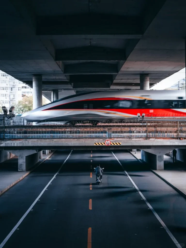 A white, red, and gray Fuxinghao high-speed train, blurred by motion, speeds across an elevated railway track. Below, a person on a motorcycle rides on a road under a concrete overpass in an urban environment.