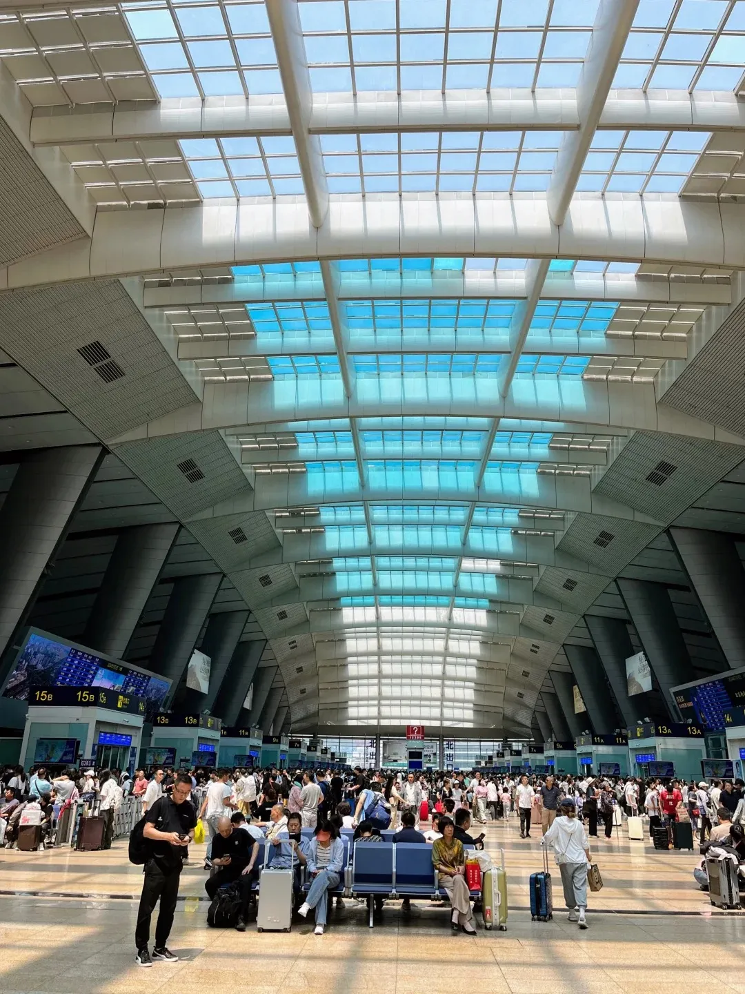 Wide interior view of a modern, bustling train station waiting hall. Hundreds of passengers with luggage are standing and sitting on blue benches under a high, skylit ceiling. The ceiling features white structural elements and numerous clear and blue-tinted glass panels. Digital display screens showing platform information like '15B' and '13A' are visible on the left.