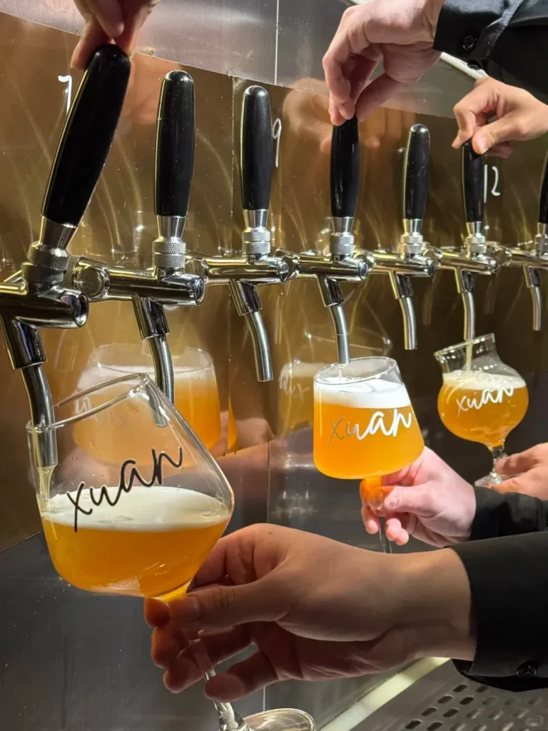 Multiple hands operating chrome beer taps against a gold-colored wall, pouring amber craft beer into various stemmed glasses. The glasses are branded with the black script 'xuan' and show a foamy head on the beer.