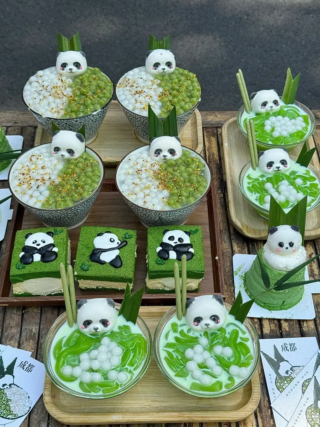 A wooden table displaying various panda-themed desserts. Four bowls contain a split serving of white tapioca pearls and green pea-like pearls, each topped with a small panda head decoration. Several glass bowls hold green liquid, green cendol noodles, and white tapioca pearls, also featuring panda heads and green bamboo stick decorations. Three rectangular green matcha cakes with white filling are topped with black and white panda figures. A small round green cake shaped like a bamboo shoot is crowned with a panda figure. Small cards with '成都' (Chengdu) in Chinese are visible.