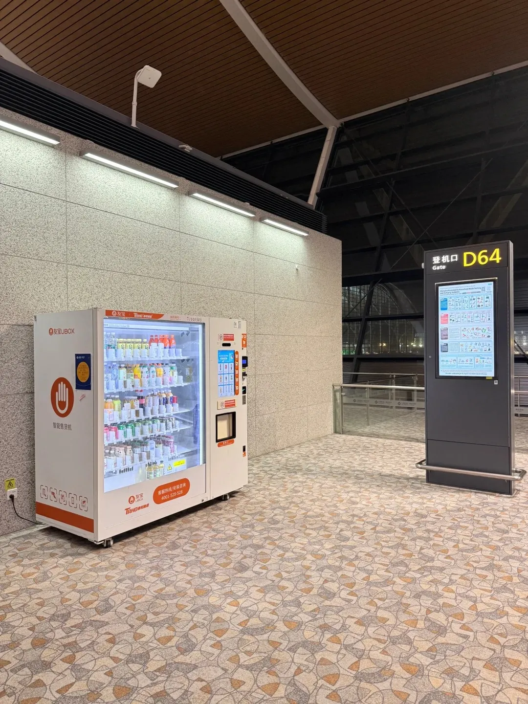 A white UBOX smart vending machine filled with various bottled drinks stands beside a black digital gate information board displaying 'Gate D64' in a modern airport terminal. The floor is covered with patterned carpet, and the ceiling has wooden panels.