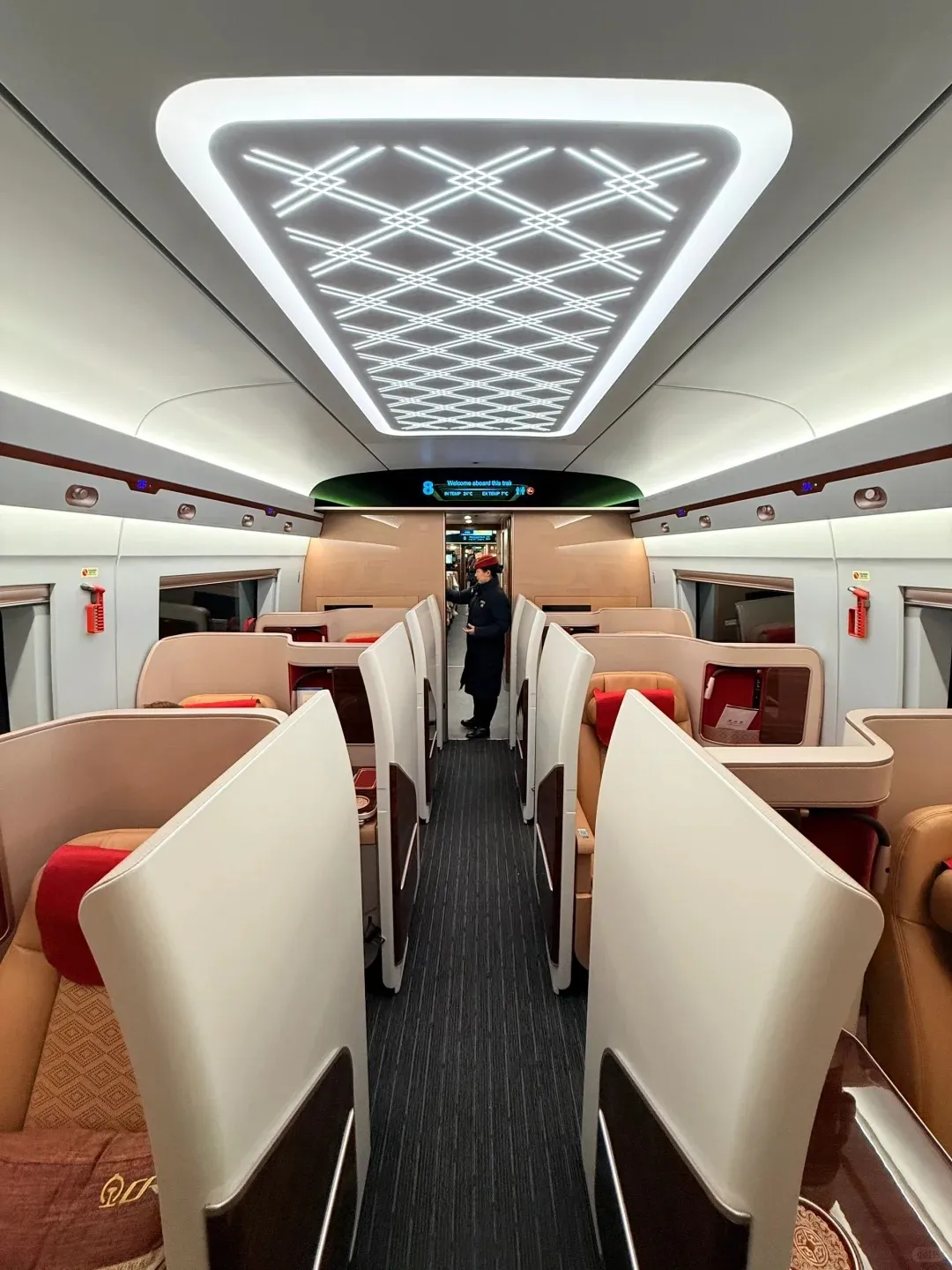 Interior of a modern high-speed train first class cabin, featuring spacious individual seats with brown and cream upholstery, red headrests, and privacy dividers. A train attendant in a dark uniform stands by the illuminated display at the end of the aisle. The ceiling features a large, patterned LED light panel.