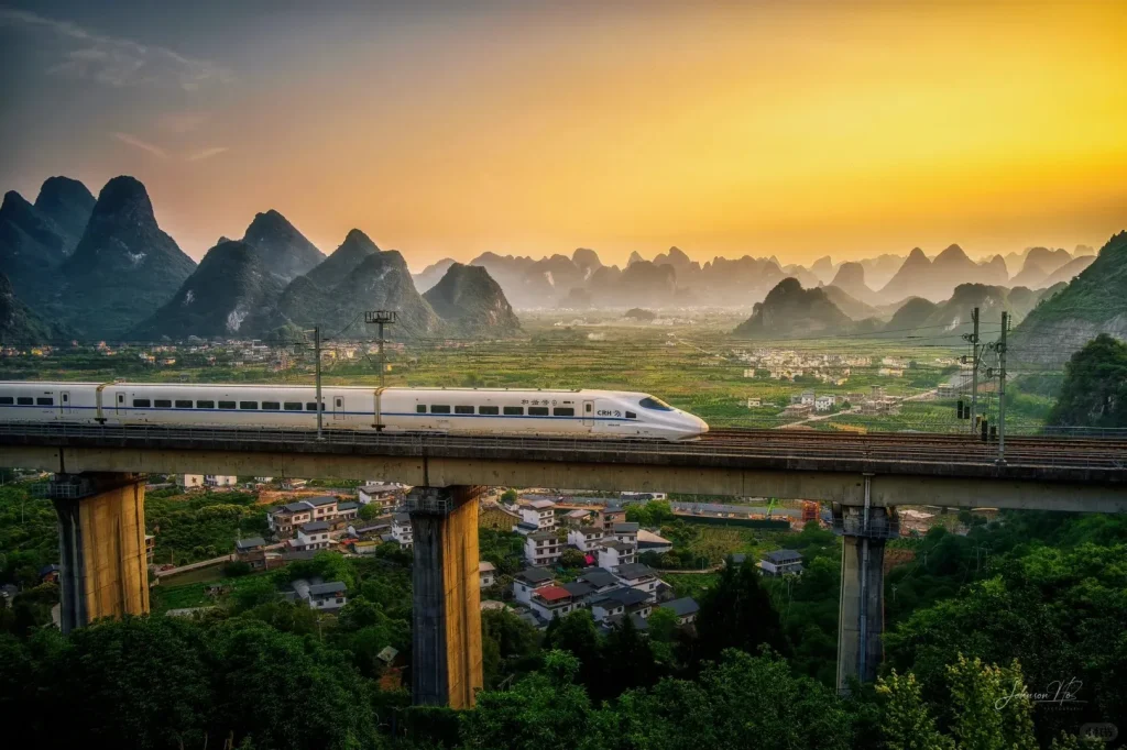 A white high-speed CRH train with blue accents travels on an elevated concrete railway bridge. Below the bridge are green trees and small village houses. In the background, numerous green, conical karst mountains stretch into the distance under a golden orange sunset sky.