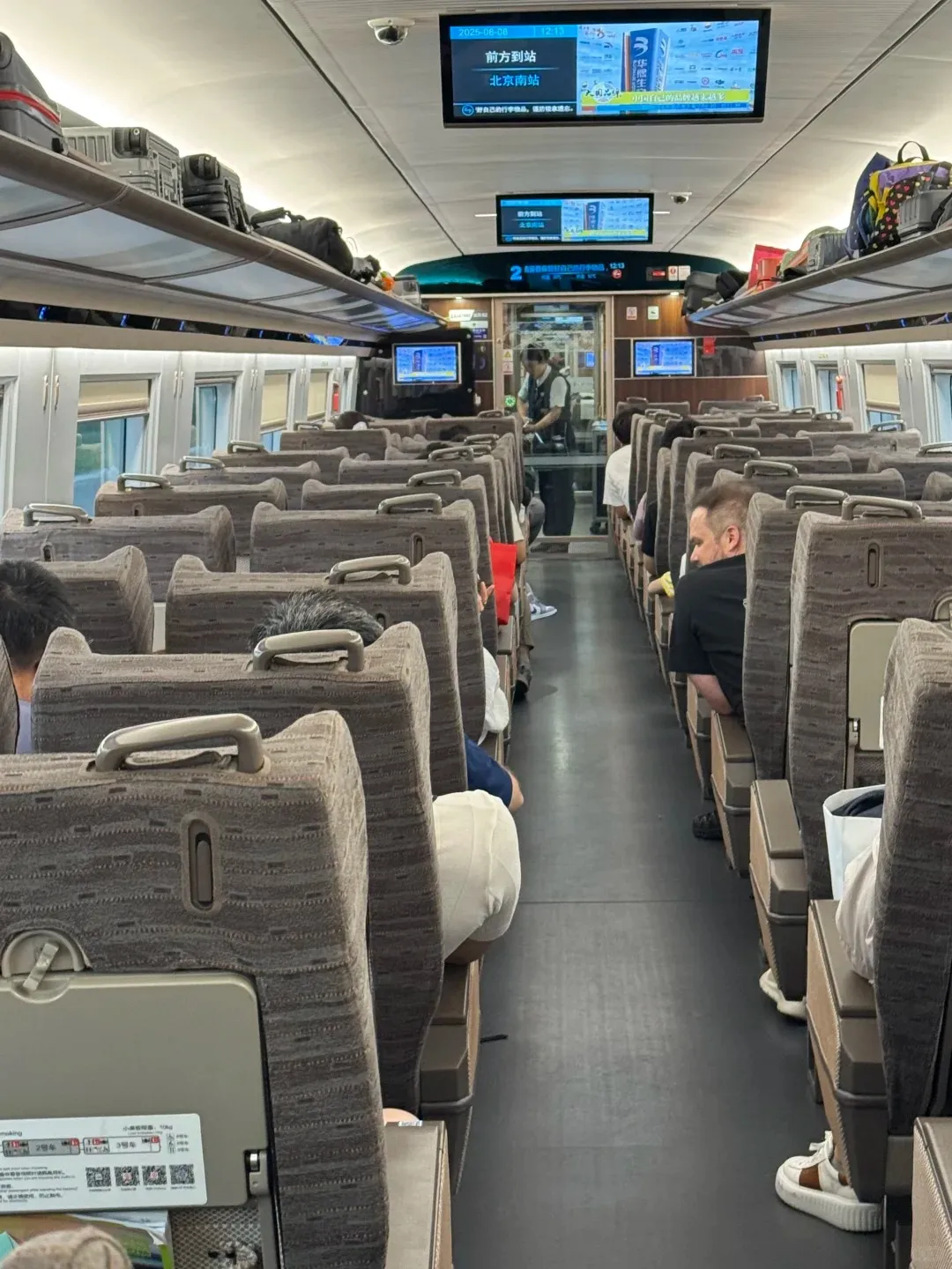 Interior view of a Chinese high-speed train carriage with rows of brown and grey fabric seats. Passengers are visible seated and walking in the aisle. Overhead screens display information in Chinese, including 'Next stop: Beijing South Station', and luggage racks above the seats are filled with bags.