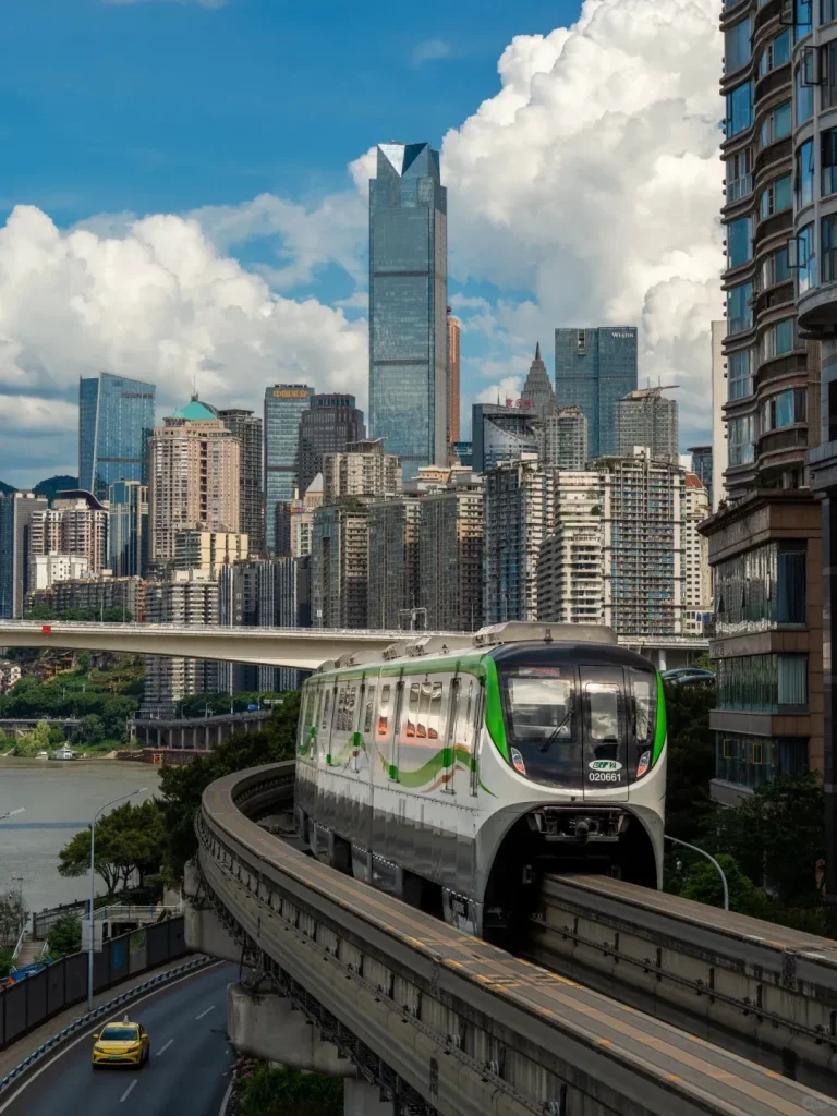 A white and green Chongqing monorail train curves on an elevated track, heading towards the viewer. In the background, a dense city skyline features many modern skyscrapers and residential buildings under a bright blue sky with large white clouds. A prominent tall building with a distinctive pointed top dominates the center of the skyline. To the left, a wide river flows, with a bridge visible in the distance, and a yellow taxi drives on a road beneath the monorail track.