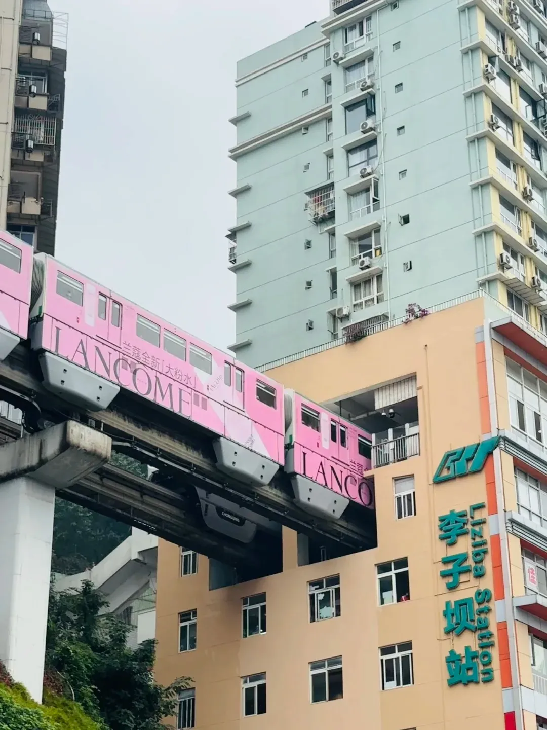 A pink Lancome-branded monorail train is seen passing directly through the multi-story Liziba Station building in Chongqing. The elevated monorail track runs through the tan-colored building, with a taller light blue residential building in the background. Green trees and foliage are visible below the track.