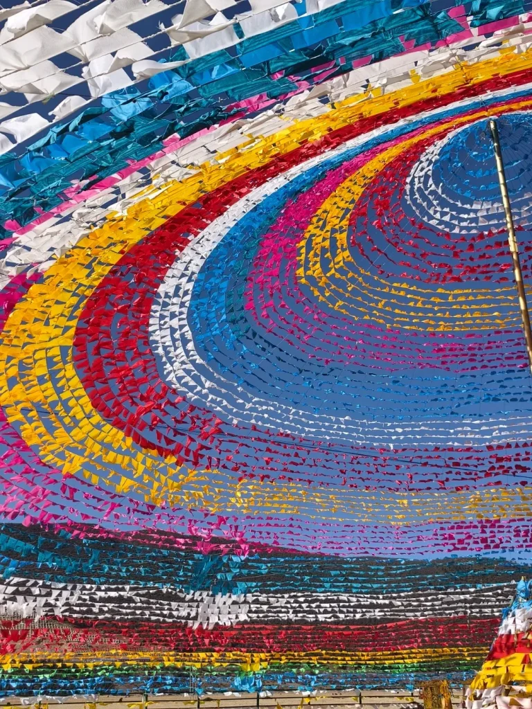 Thousands of small, brightly colored triangular flags strung across a clear blue sky. The flags are arranged in vibrant swirling patterns of white, blue, yellow, red, pink, and black, forming a dense, decorative canopy.