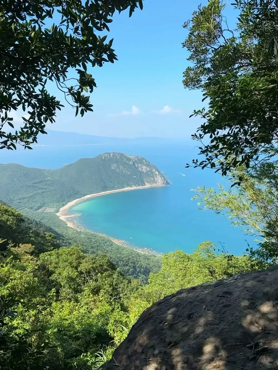 High-angle view of a scenic bay with a long curving sandy beach and a forested peninsula extending into clear turquoise-blue ocean water. Lush green trees frame the view from above, with a large rock in the bottom right foreground. Faint hills are on the distant horizon under a bright blue sky.