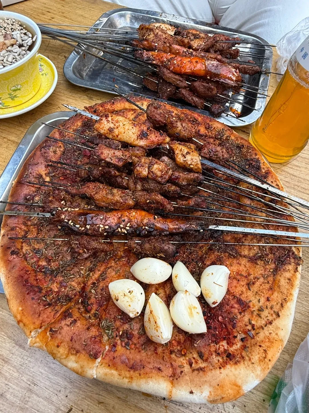 A large, round flatbread seasoned with red spices and green herbs is covered with many metal skewers of grilled meat and some seafood. Six peeled garlic cloves sit on the bread. In the upper background, another tray of grilled skewers, a bottle of beer, and a yellow bowl of seeds are visible on a wooden table.