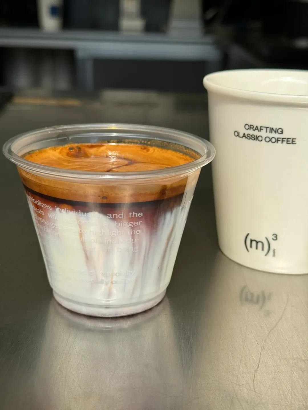 A clear plastic cup displays a layered dirty coffee with a thick, dark espresso crema on top, separated from a white milk base, with dark streaks subtly mixing into the milk. To its right, a white ceramic cup reads 'CRAFTING CLASSIC COFFEE' and features the logo '(m)³₁'. Both cups sit on a reflective, brushed metal surface.