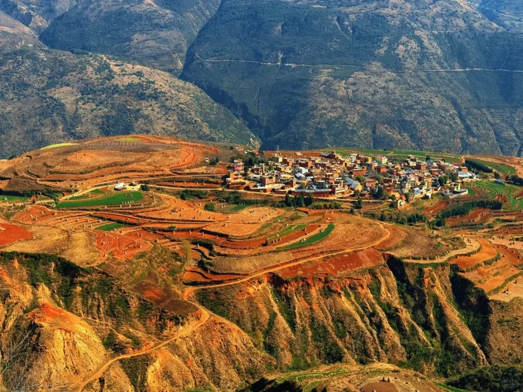 An expansive aerial view of the Dongchuan Red Land, showing a village situated amidst rolling hills with striking red soil terraced fields and patches of green crops. Distant mountains rise in the background under a blue sky, and winding dirt roads traverse the landscape.