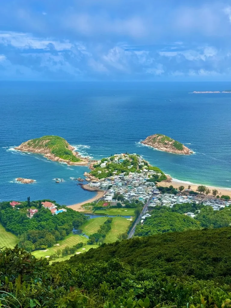 Elevated panoramic view of Hong Kong's Shek O peninsula, featuring a coastal village with numerous buildings, two sandy beaches, a lush green golf course, and verdant hills. Several small rocky islands are visible in the deep blue ocean under a partly cloudy sky.