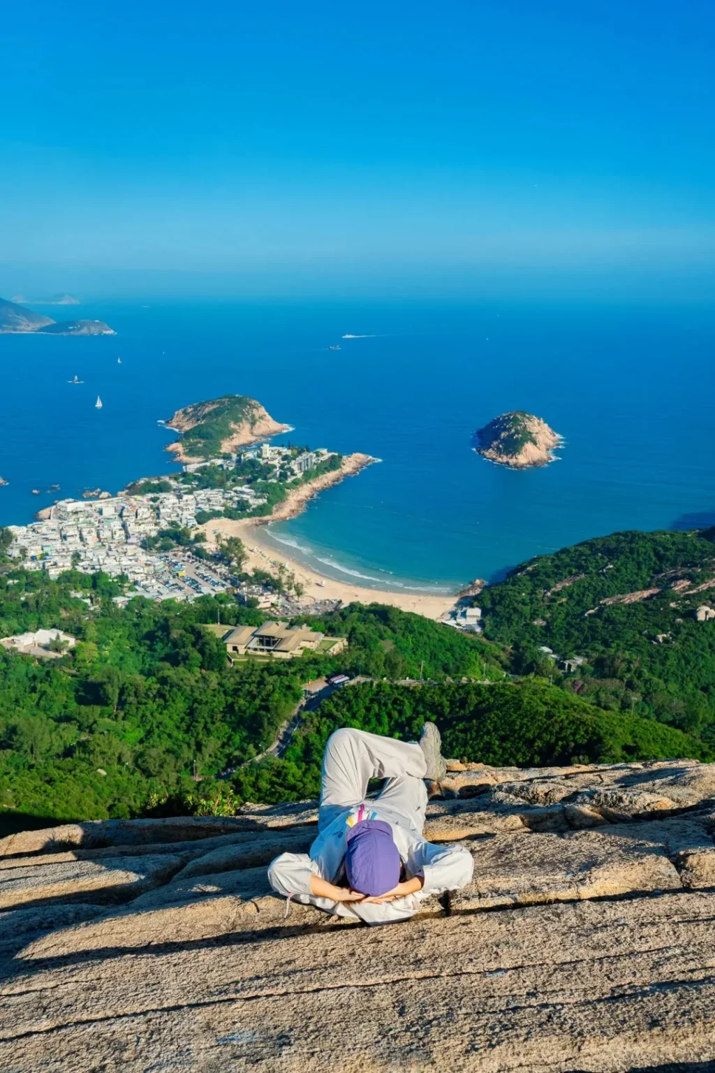 A person wearing a light grey jacket, light grey pants, and a purple cap lies on their back on a large, textured grey rock, with hands behind their head. They are facing away from the camera, looking down at a vast panoramic coastal view. The view includes lush green hills, a winding road, the white and light-colored buildings of Shek O village, a curved sandy beach, and the deep blue ocean. In the ocean are several small, rocky islands and a few boats. The horizon shows distant land under a bright clear blue sky.