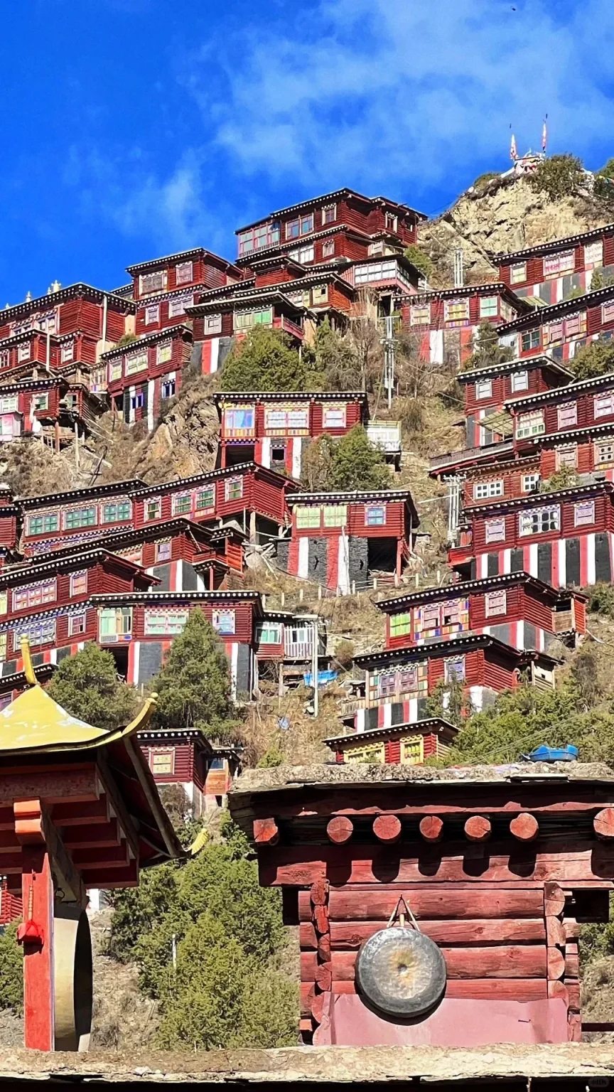 Numerous red wooden monastic dwellings with colorful window frames, some with vertical red, white, and black stripes, built into a rocky green hillside under a bright blue sky. In the foreground, parts of a traditional yellow-roofed building and a red wooden structure with a circular metal gong hanging on its side are visible.