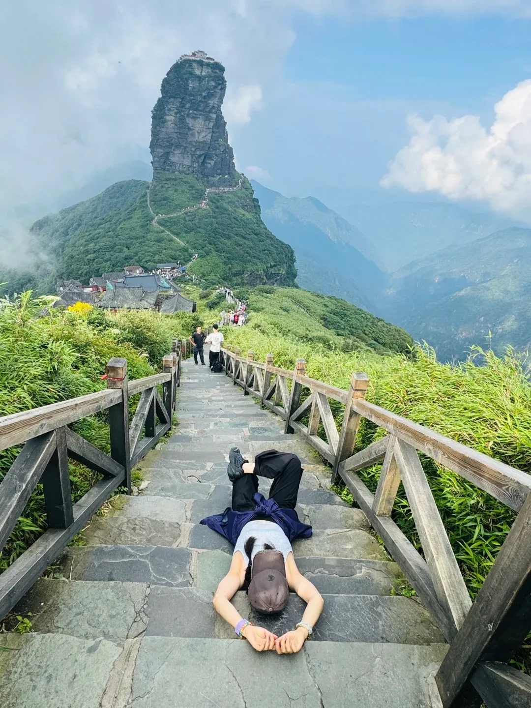 A person lies face down on a grey stone staircase with wooden railings, taking a rest. In the background, the towering, narrow Fanjingshan Golden Summit rises dramatically, featuring a temple complex on its peak, surrounded by winding paths, lush green mountains, and a partly cloudy sky.