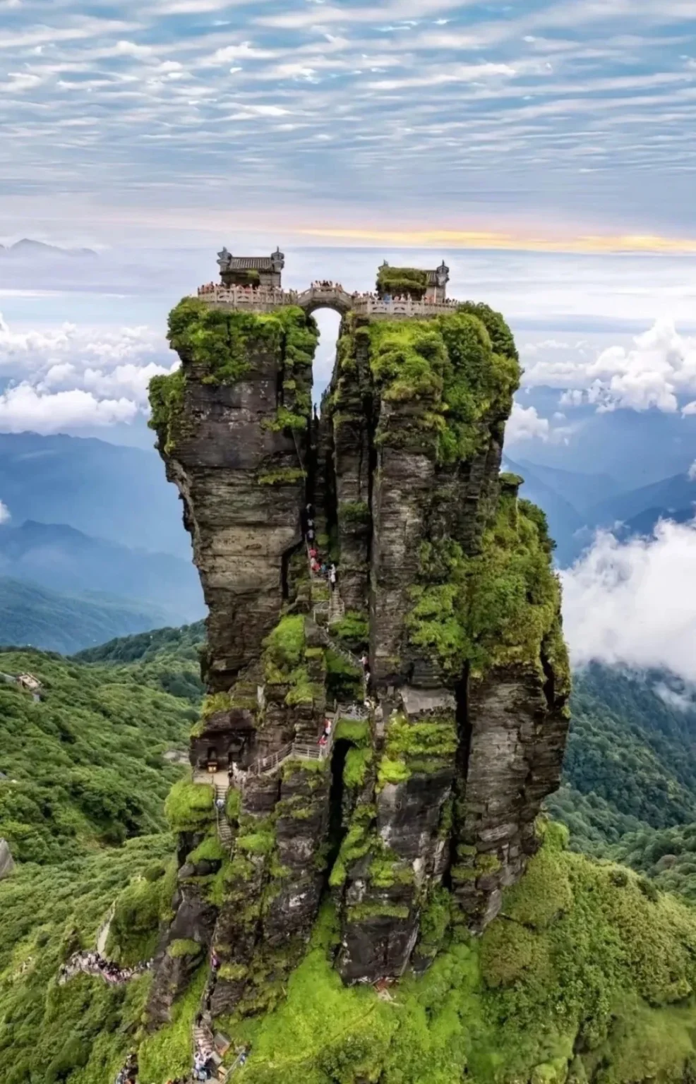 Fanjingshan Red Clouds Golden Summit featuring two steep, moss-covered rock pinnacles. A stone bridge connects two traditional temples perched atop the peaks. Winding staircases with hikers are built into the rock face, surrounded by lush green mountains and clouds.