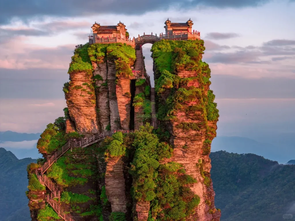 Two ornate Chinese temple buildings with dark roofs stand on separate, twin peaks of a tall, narrow mountain, connected by a small stone arch bridge. Winding wooden staircases are built into the reddish-brown, moss-covered rock faces, leading up to the summits. Distant green mountains are visible in the background under a partly cloudy sky with pink and blue hues.