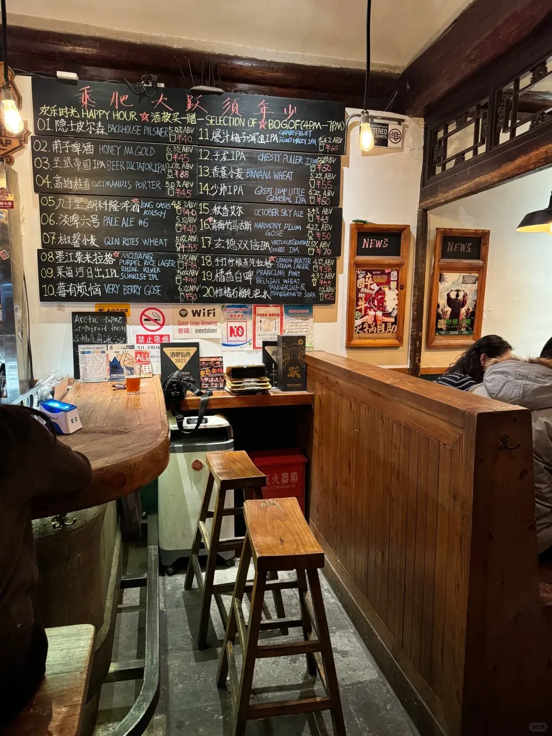 Interior view of a rustic craft beer bar featuring a large blackboard menu with beer selections and prices written in Chinese. A curved wooden bar counter is visible with two empty wooden high stools in the foreground and a person seated at the far end. The walls are made of traditional wooden paneling, and two framed 'NEWS' signs hang on the right wall. Warm pendant lights illuminate the space.