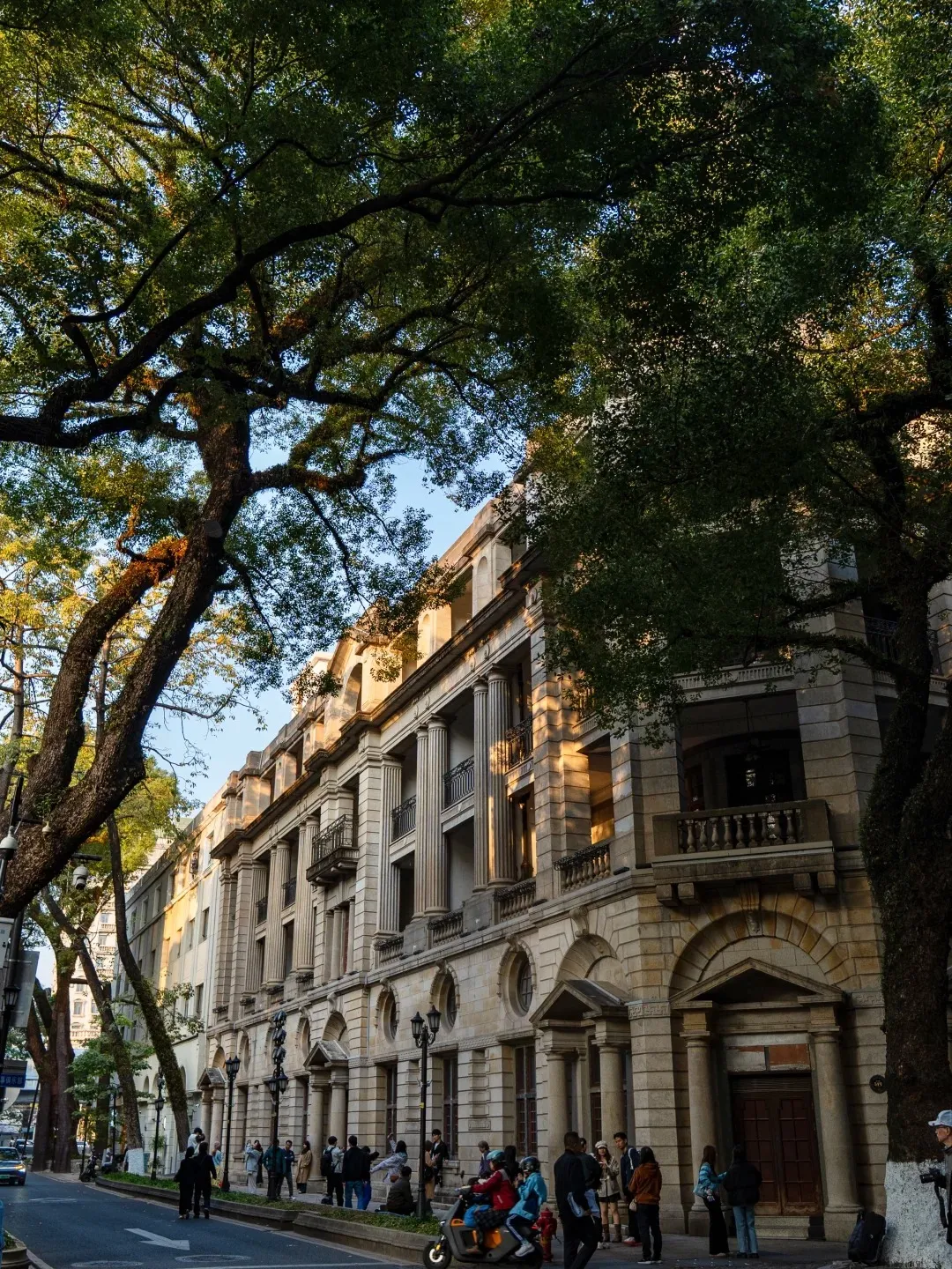 A grand colonial-style stone building with multiple stories, classical columns, and ornate balconies lines a street. Large banyan trees with extensive green canopies frame the building from above and the left. Warm sunlight illuminates parts of the building facade and filters through the tree leaves. Several people are walking on the sidewalk in front of the building.