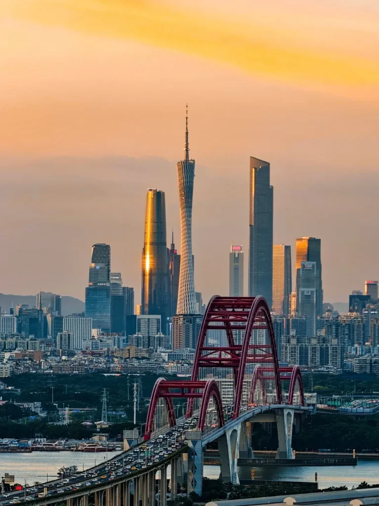 Panoramic view of Guangzhou cityscape at sunset, featuring the slender Canton Tower and numerous modern skyscrapers reflecting golden light. In the foreground, a multi-arched red bridge crosses the Pearl River, bustling with traffic.