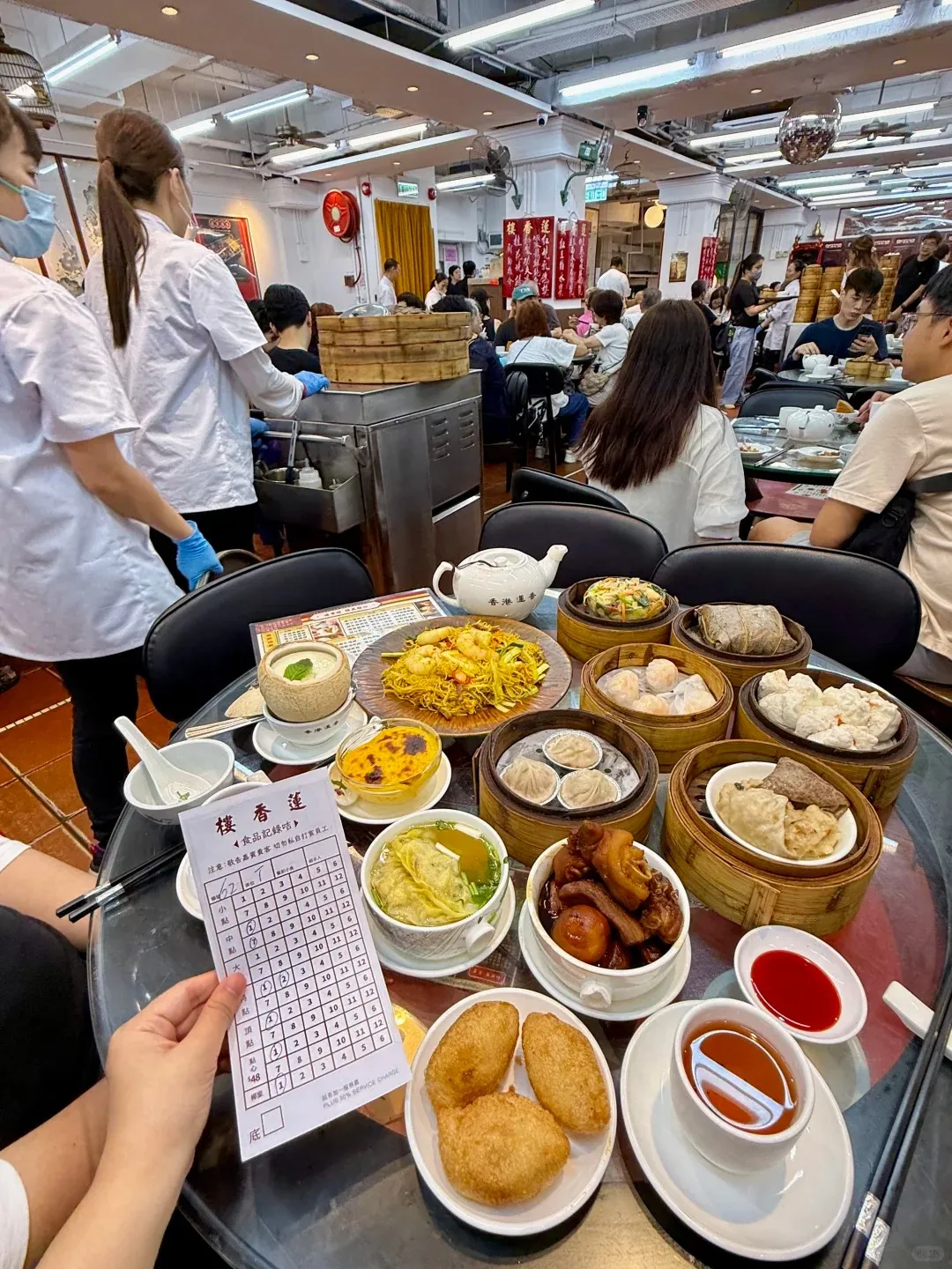 A bustling, brightly lit traditional Cantonese dim sum restaurant. In the foreground, a round table is covered with many small dishes including bamboo steamers filled with various dumplings, a plate of fried pastries, a bowl of noodles, braised meats, a teapot, and a paper order form. People are dining at tables throughout the restaurant, and waitstaff in white uniforms push dim sum trolleys in the background.