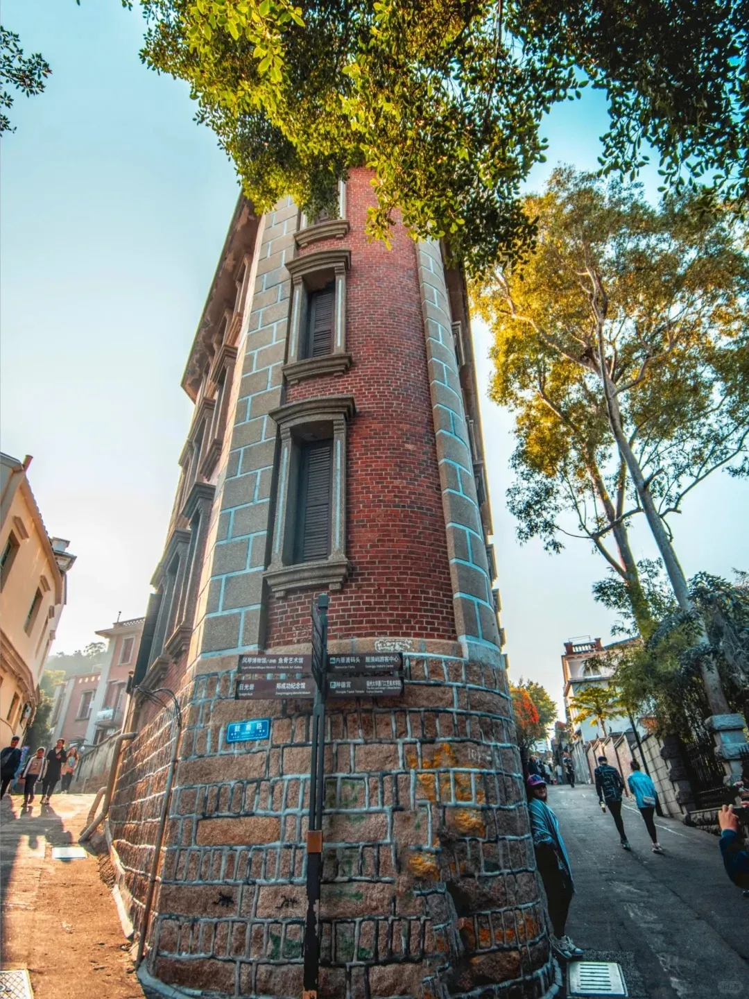 A narrow pedestrian street on Gulangyu Island featuring a colonial-style corner building with a rough stone block base and red brick upper floors. Green trees with dense foliage are visible against a bright blue sky. Several pedestrians walk along the street, and a street sign with Chinese characters stands at the corner.