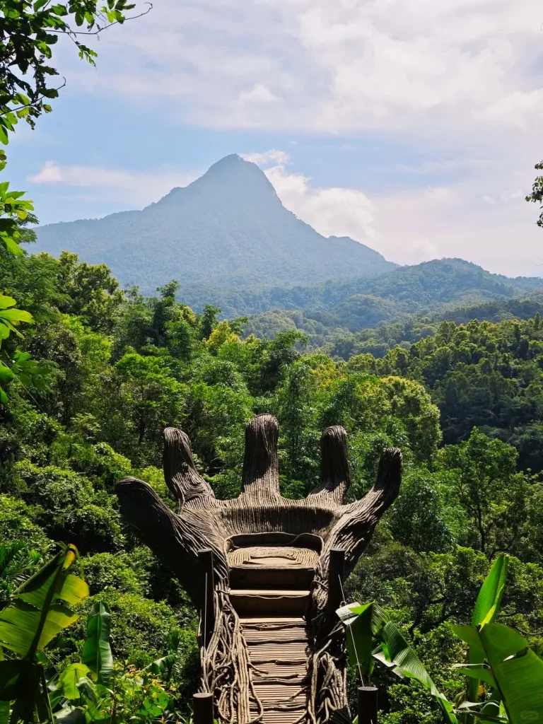 A large wooden hand-shaped viewing platform with steps leading to a seating area, surrounded by lush green tropical foliage. In the background, the prominent, jagged peak of Wuzhishan Five Finger Mountain rises above a dense tropical rainforest under a partly cloudy blue sky.