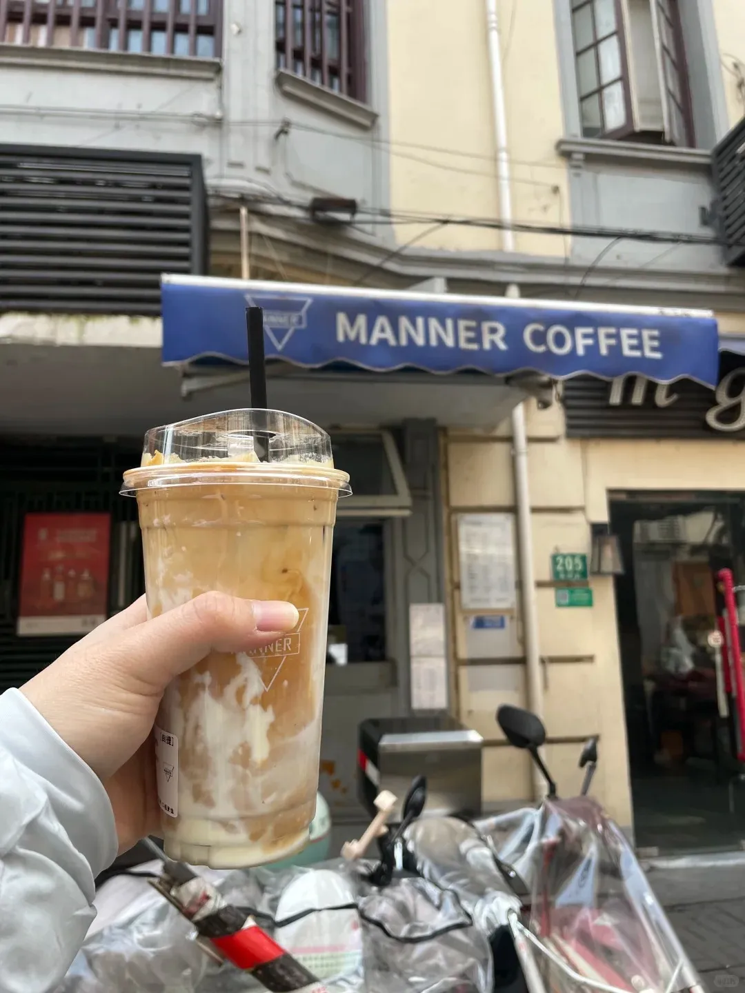 A person's hand holds a clear plastic cup of layered iced coffee with a black straw, positioned in front of a Manner Coffee shop. The shop features a blue awning with 'MANNER COFFEE' in white letters. Buildings and a covered scooter are visible in the street background.