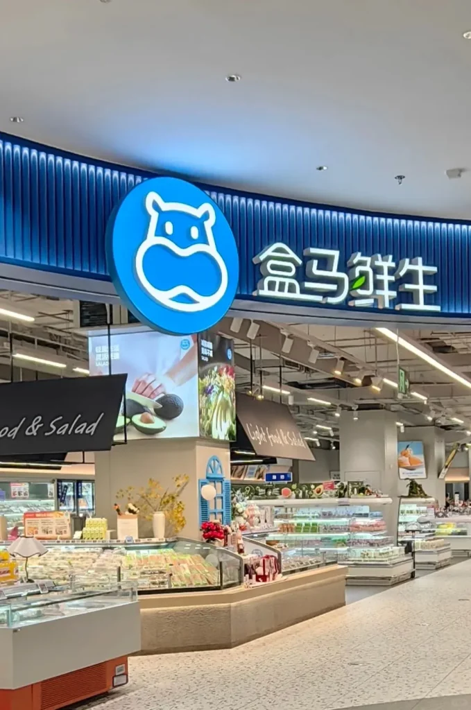 Interior view of a modern Hema Freshippo supermarket. A prominent blue sign features a white hippo logo and the Chinese characters '盒马鲜生' (Hema Xiansheng). Below the sign, multiple glass display cases are filled with various packaged fresh foods, salads, and desserts. Signs overhead read 'Food & Salad' and 'Light Food & Salad'.