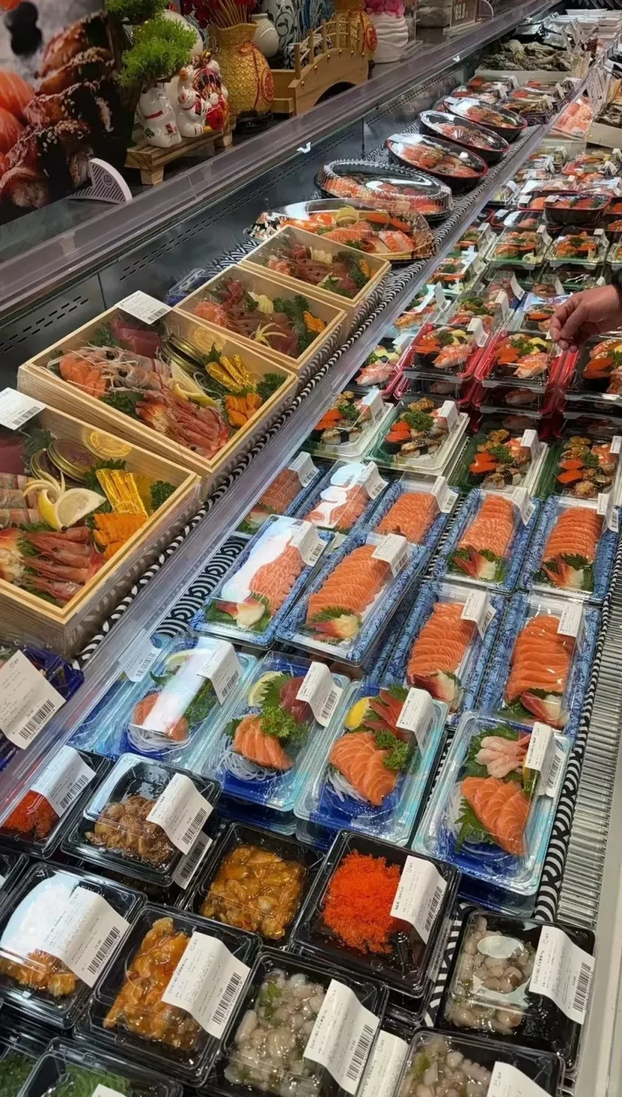 A long, refrigerated supermarket display case filled with numerous pre-packaged Japanese food items. The display features rows of clear plastic containers holding slices of salmon sashimi, various sushi rolls, nigiri, and small seafood salads like octopus and tobiko. Several wooden bento-style boxes are also visible, packed with assorted sashimi including salmon, tuna, and shrimp, garnished with lemon slices and greens. A person's hand is visible towards the right, reaching for an item.
