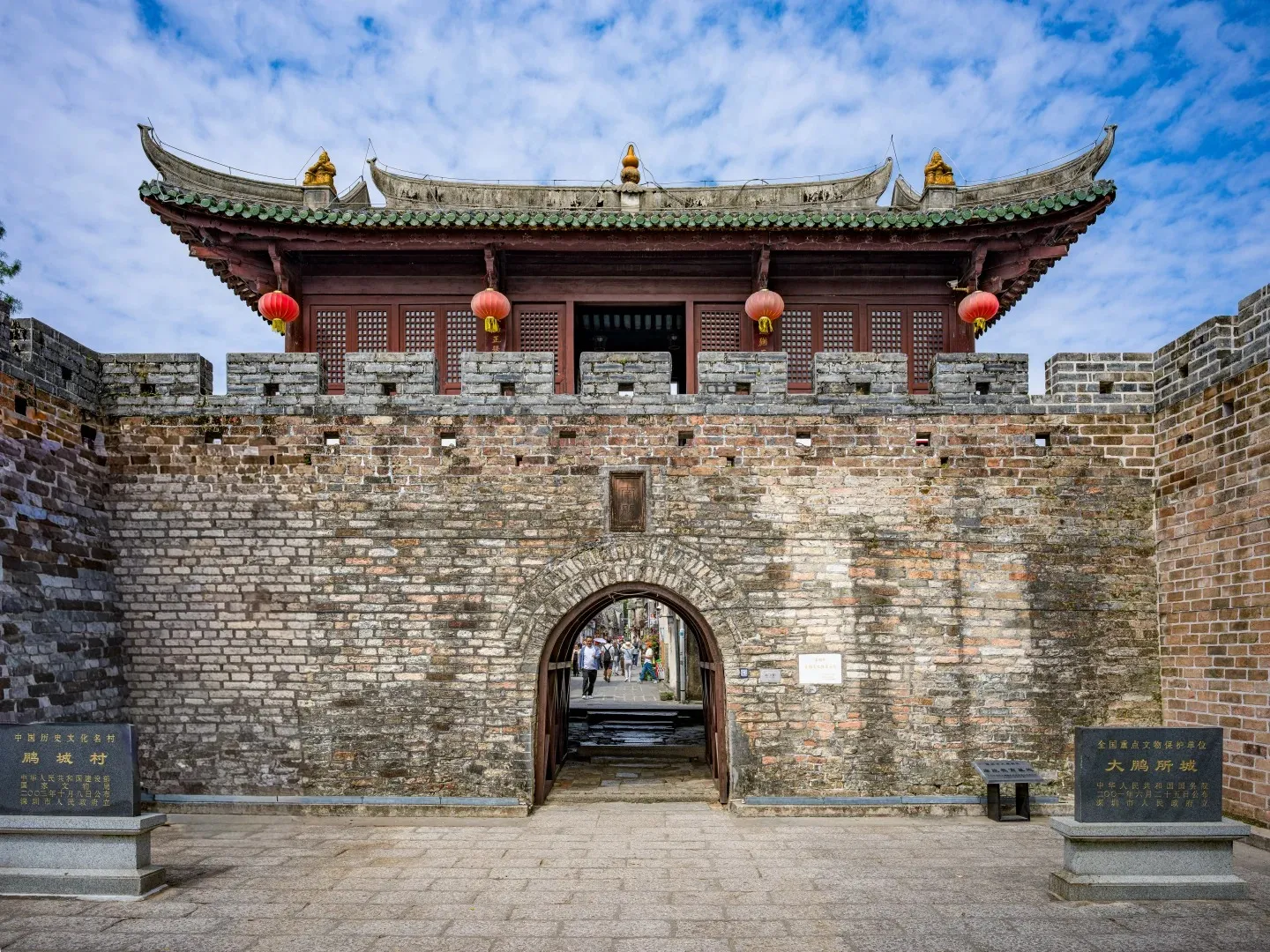 A large, weathered brick fortress wall with a central arched wooden gateway. Above the wall, a traditional Chinese building with a green tiled roof, red wooden elements, and four red lanterns hangs. Two dark stone information plaques stand on the paved ground in front of the wall.