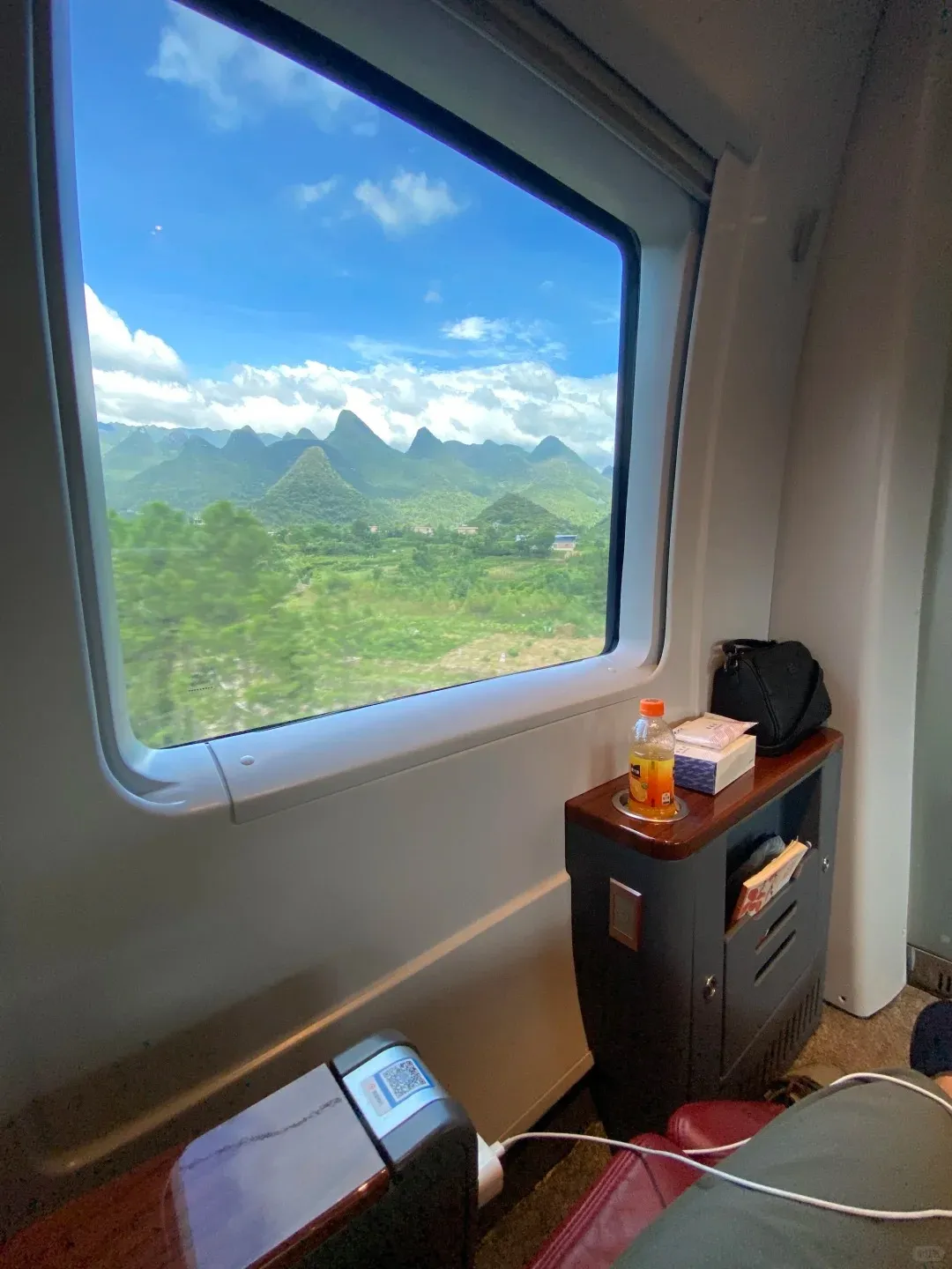 Interior view from a high-speed train window showing lush green karst mountains and valleys under a blue sky with white clouds. Inside the train, a wooden-topped side table with a black bag, an orange drink, and a snack box sits next to the window. A charging cable is plugged into a seat armrest in the foreground.