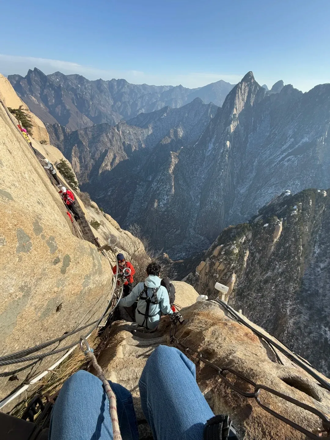A first-person view from a hiker's perspective shows legs in blue jeans and a safety harness looking down a steep, narrow path on the side of Mount Hua. Ahead, several other hikers navigate the wooden plank walk, secured by thick metal chains and carabiners, with a vast valley of rugged, snow-dusted mountains stretching into the distance under a clear blue sky.