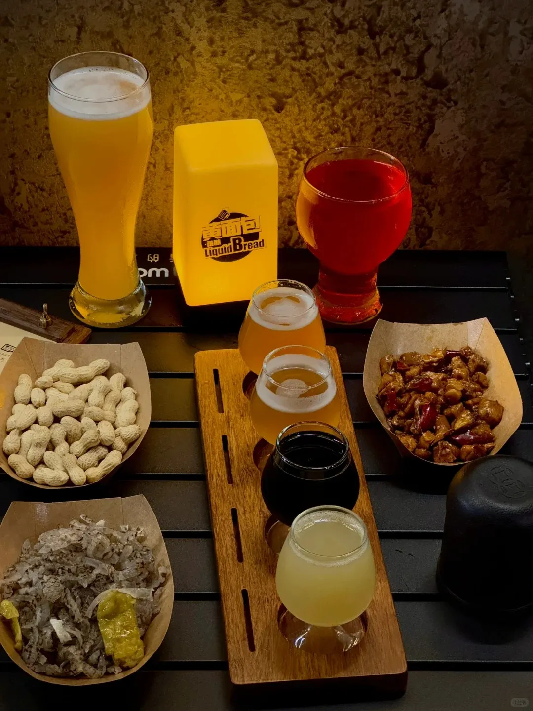 A tabletop view featuring a craft beer tasting flight on a wooden board with three small glasses (a light golden beer, a dark brown beer, and a hazy pale yellow beer), alongside a tall pilsner glass of golden beer and a red beverage. Three paper bowls contain different snacks: shelled peanuts, a shredded meat dish with white onions and yellow peppers, and a spicy dish with dark chunks and red chilies. A glowing yellow rectangular lamp displaying the 'Liquid Bread' logo is visible in the background.