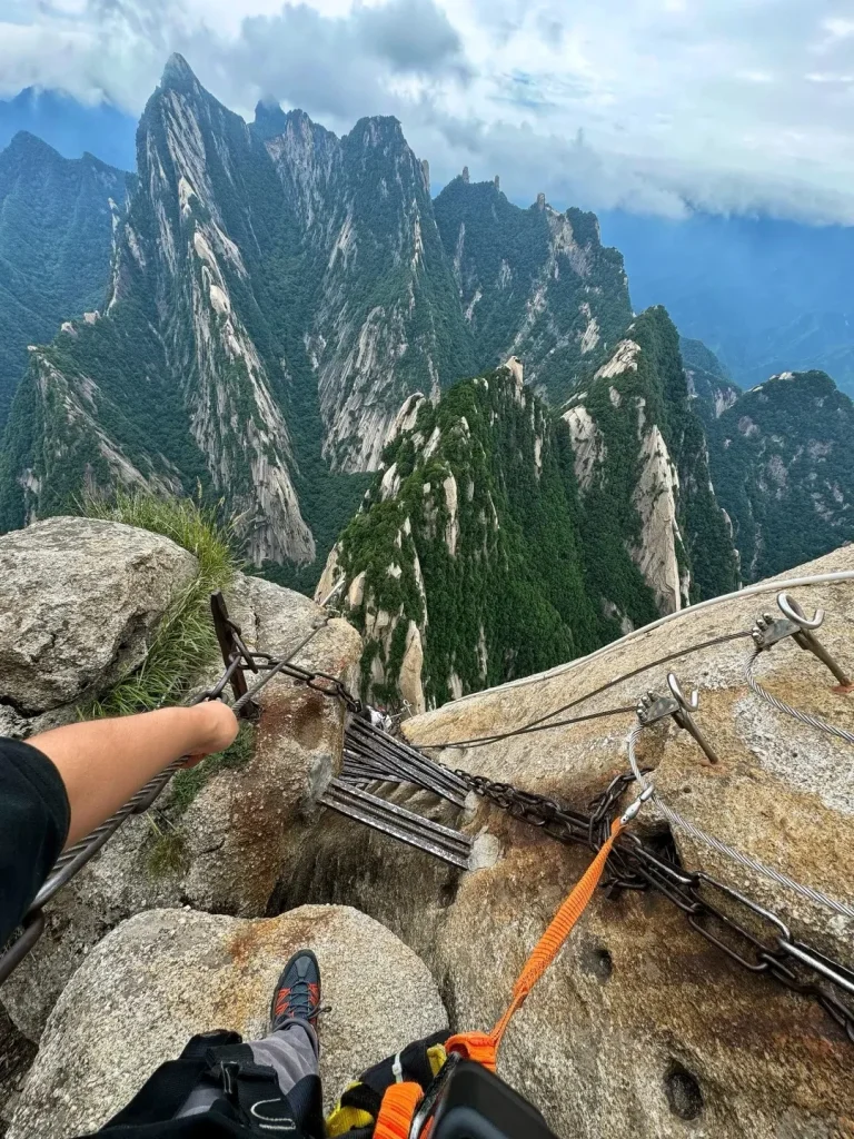 A first-person view showing a person's arm holding a steel safety cable and their foot on a rocky mountain path. An orange safety lanyard from a harness is clipped to a thick metal chain secured to the rock. Ahead, a narrow path constructed from horizontal metal beams is built into the side of a steep cliff. In the background, dramatic, rugged mountains covered in green trees rise under a cloudy sky.
