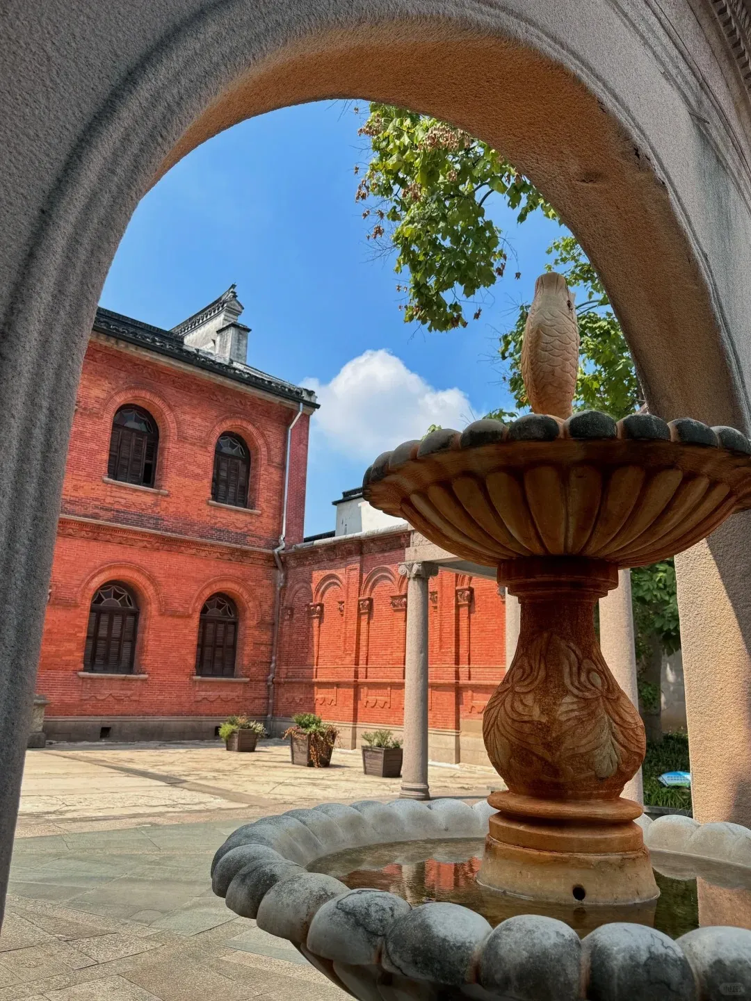 View through a grey stone archway of a two-story red brick building with dark arched windows and shutters. In the foreground, a multi-tiered brown and grey stone fountain featuring a carved fish sculpture on its top tier is visible. The scene includes a paved courtyard with potted plants, partially visible stone columns, a bright blue sky with white clouds, and green tree branches.