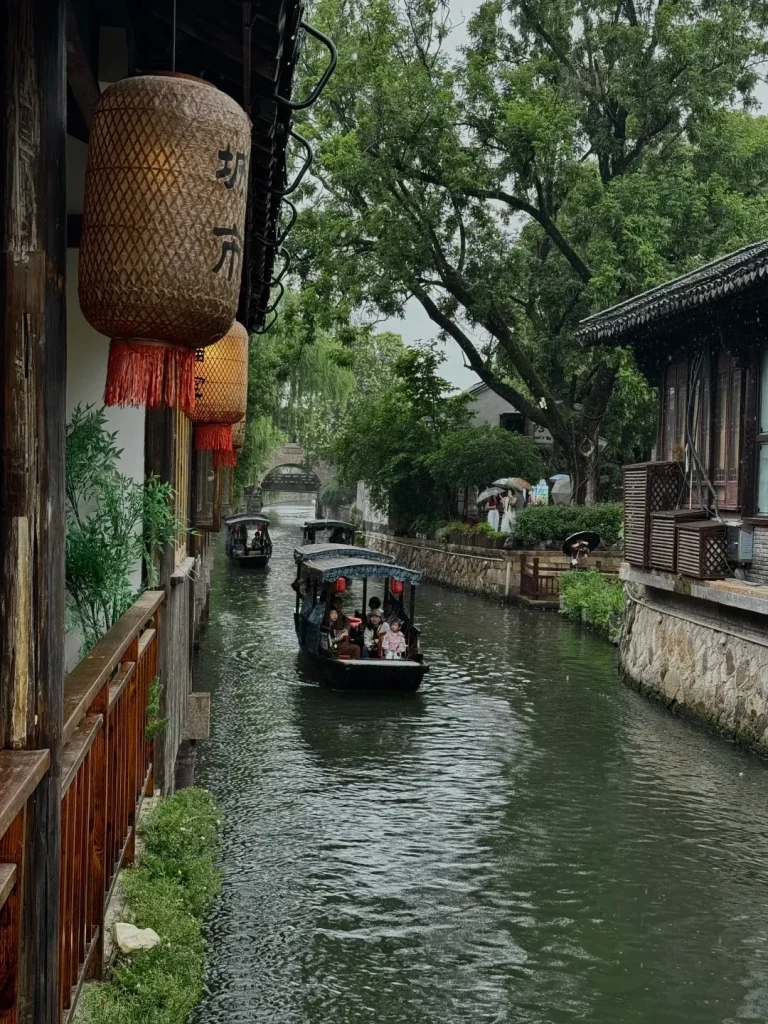 A narrow canal in Nanxun Water Town with three dark wooden boats carrying passengers. On the left, a historic wooden building features two large woven lanterns with black calligraphy and red tassels. On the right, an old building with stone walls and dark wooden trim lines the canal. Lush green trees and a distant stone arch bridge are visible in the background.