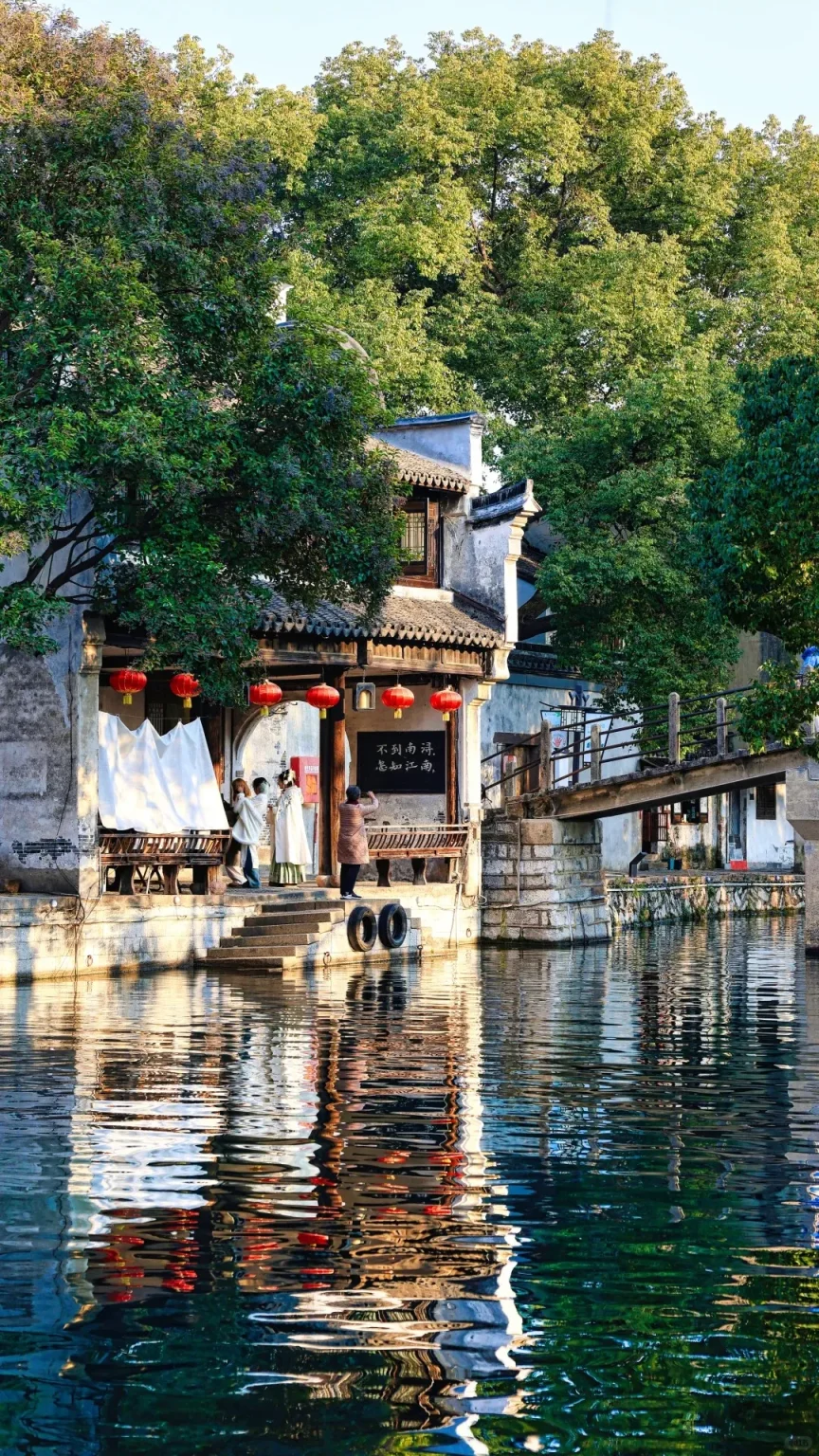 Traditional Chinese buildings with white walls, dark tiled roofs, and red lanterns line a calm canal in Nanxun Water Town. On a waterside platform, two people in traditional Hanfu-style clothing pose while another person photographs them. Stone steps descend to the water next to old tire bumpers. A rustic wooden bridge crosses the canal in the background, and green trees canopy the scene above, with reflections visible in the water.