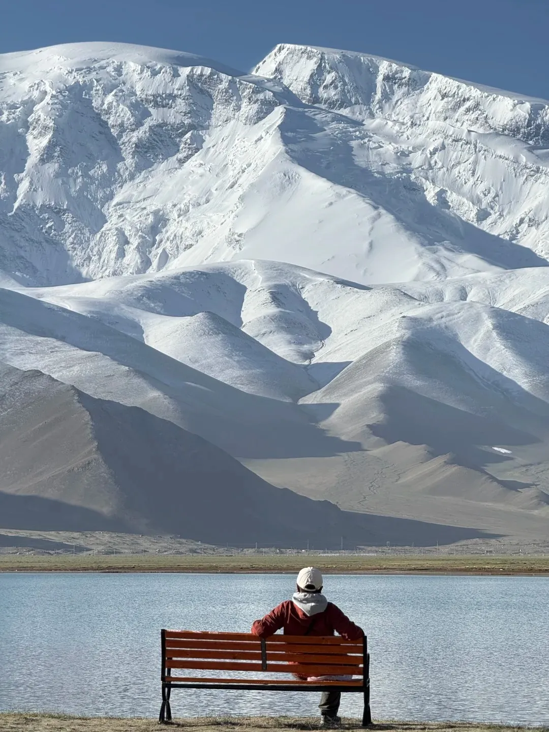 A person in a red jacket and white baseball cap sits on a wooden bench, looking out at the calm, blue waters of Karakul Lake. Beyond the lake, the massive, snow-covered peaks of Muztagh Ata mountain rise dramatically under a clear blue sky, with lighter snow-dusted foothills leading down to the shore.