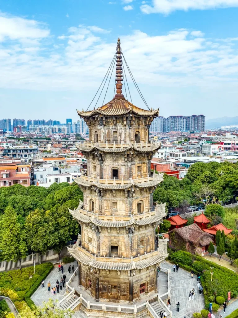 An ancient, multi-story octagonal stone pagoda from Kaiyuan Temple in Quanzhou, Fujian province, featuring intricate carvings, multiple tiered roofs with upturned eaves, and a spired finial at its peak. The pagoda is surrounded by lush green trees and landscaped pathways with people walking, set against a backdrop of a modern city skyline under a partly cloudy blue sky.