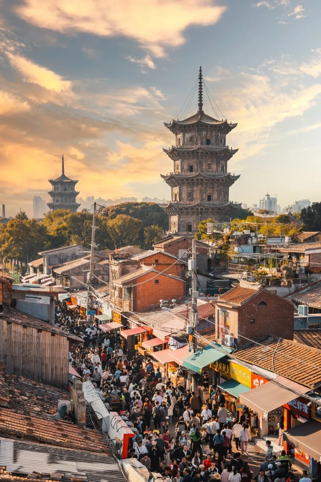An elevated view of a bustling street market in Quanzhou, China, at sunset. Two ancient stone pagodas, identified as the Kaiyuan Temple Twin Pagodas, rise prominently above a dense cluster of traditional buildings and green trees. The narrow market street below is packed with people walking between numerous vendor stalls with colorful awnings, all bathed in warm golden light under a sky with scattered orange clouds.