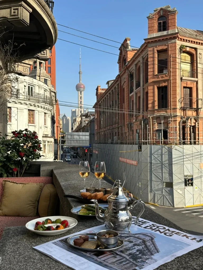 A table on an outdoor balcony featuring two glasses of white wine, a silver teapot, small plates of snacks including fruit, nuts, cookies, and asparagus. In the background, historic European-style buildings and construction scaffolding line a street, with the Oriental Pearl TV Tower visible in the distance.