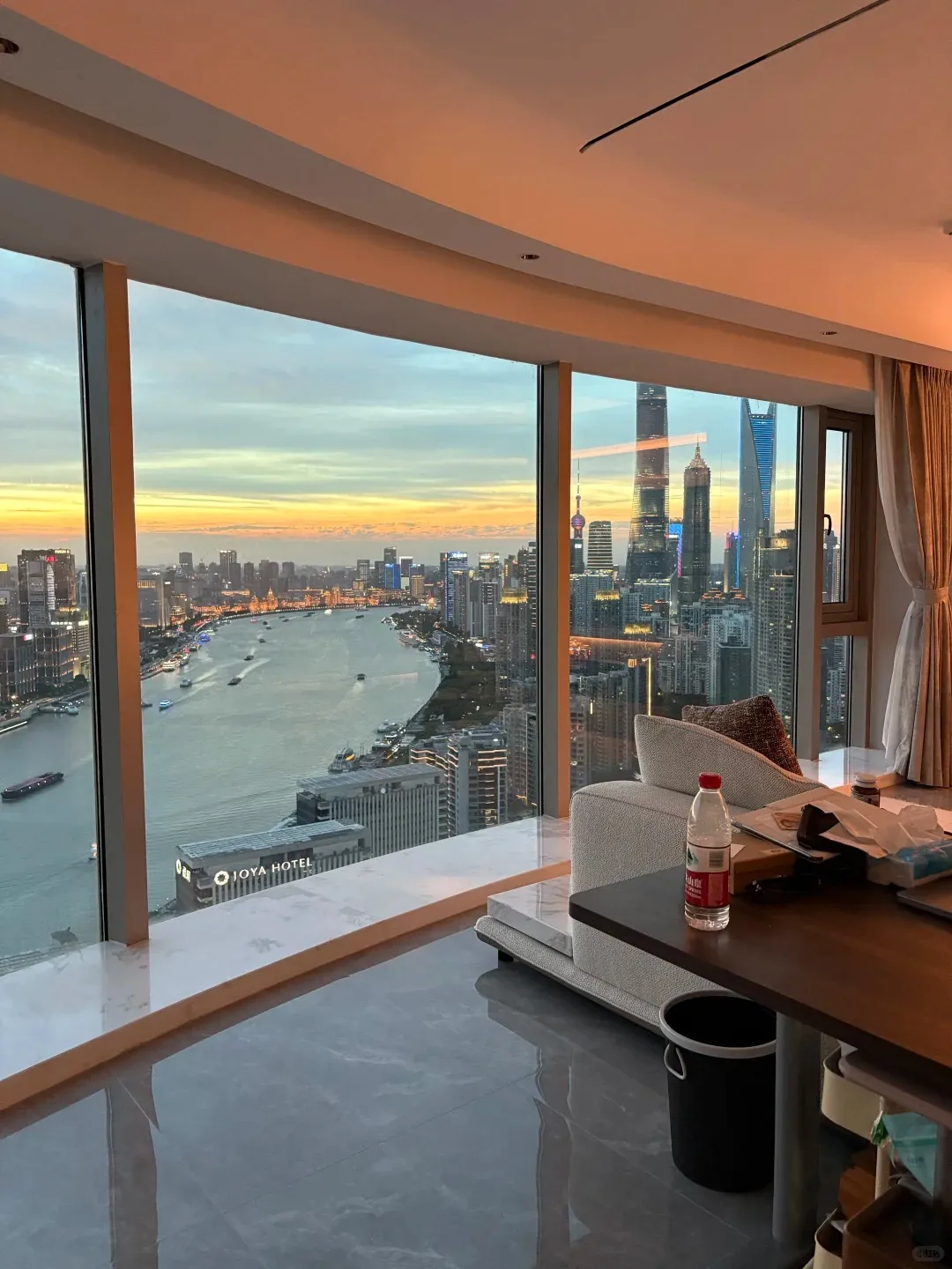 Interior view from a modern hotel room with floor-to-ceiling windows showing the Shanghai Pudong skyline, Huangpu River with boats, and city buildings at sunset. The sky is orange and blue, with prominent skyscrapers like the Shanghai Tower. A sofa and table with a water bottle are visible in the foreground.