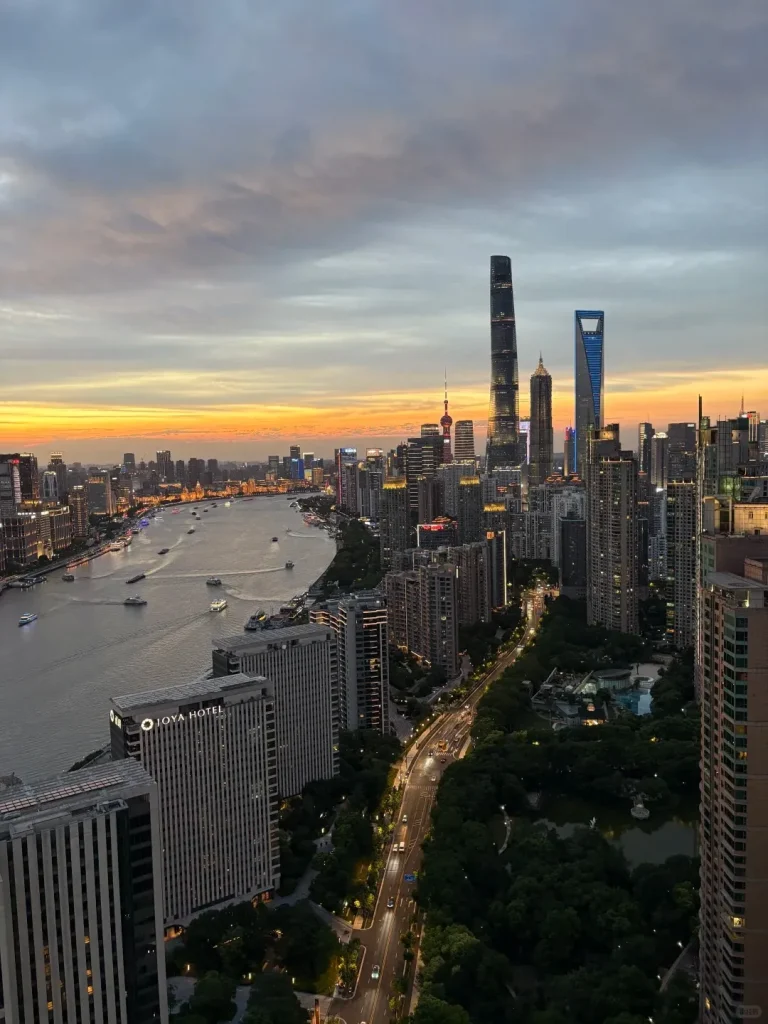 High-angle view of the Shanghai skyline at sunset, showing the Huangpu River winding through the city. On the far side, iconic Pudong skyscrapers including the Shanghai Tower, Shanghai World Financial Center, and Jin Mao Tower are brightly lit against an orange and grey sky. Buildings and roads on the near side are also illuminated, with trees and a park visible in the foreground.