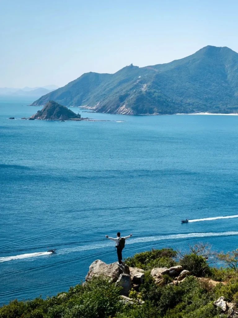 A person with a backpack and outstretched arms stands on a rocky cliff, looking out over a wide blue sea. In the background, lush green mountains and smaller islands emerge from the water, with a distant sandy beach. Two speedboats cut across the water, leaving white wakes.