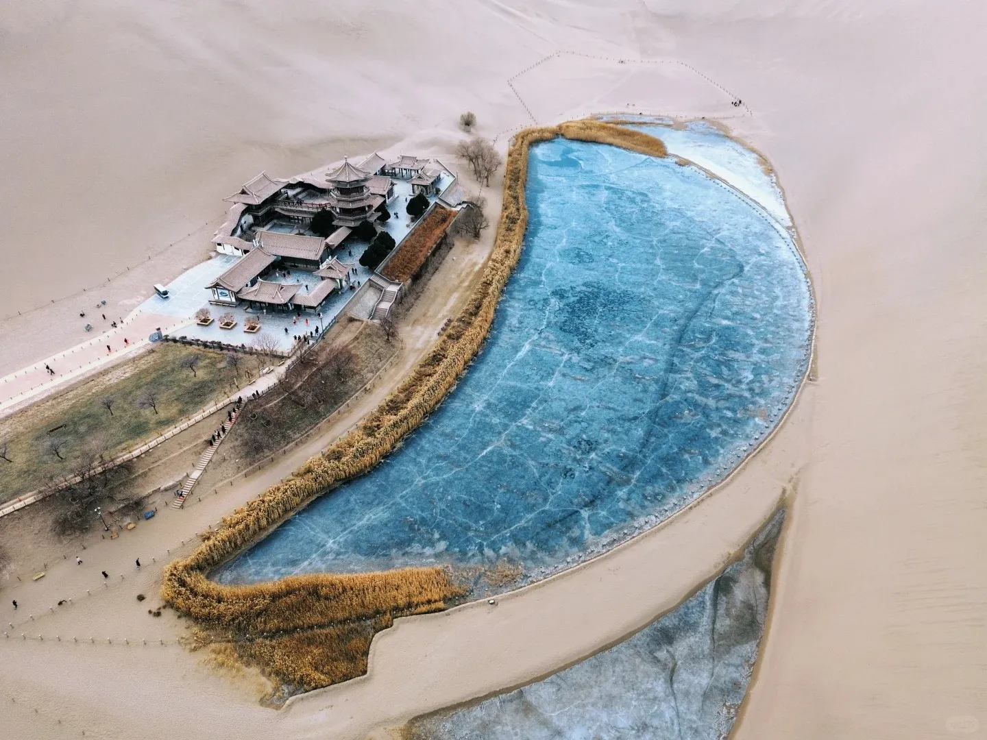 Aerial view of Dunhuang's Crescent Lake (Yueyaquan) frozen with blue ice, bordered by light brown sand dunes. Traditional Chinese pavilions with multiple tiered roofs are situated on a platform beside the lake, connected by pathways where people are walking. Dry reeds line parts of the lake's edge.