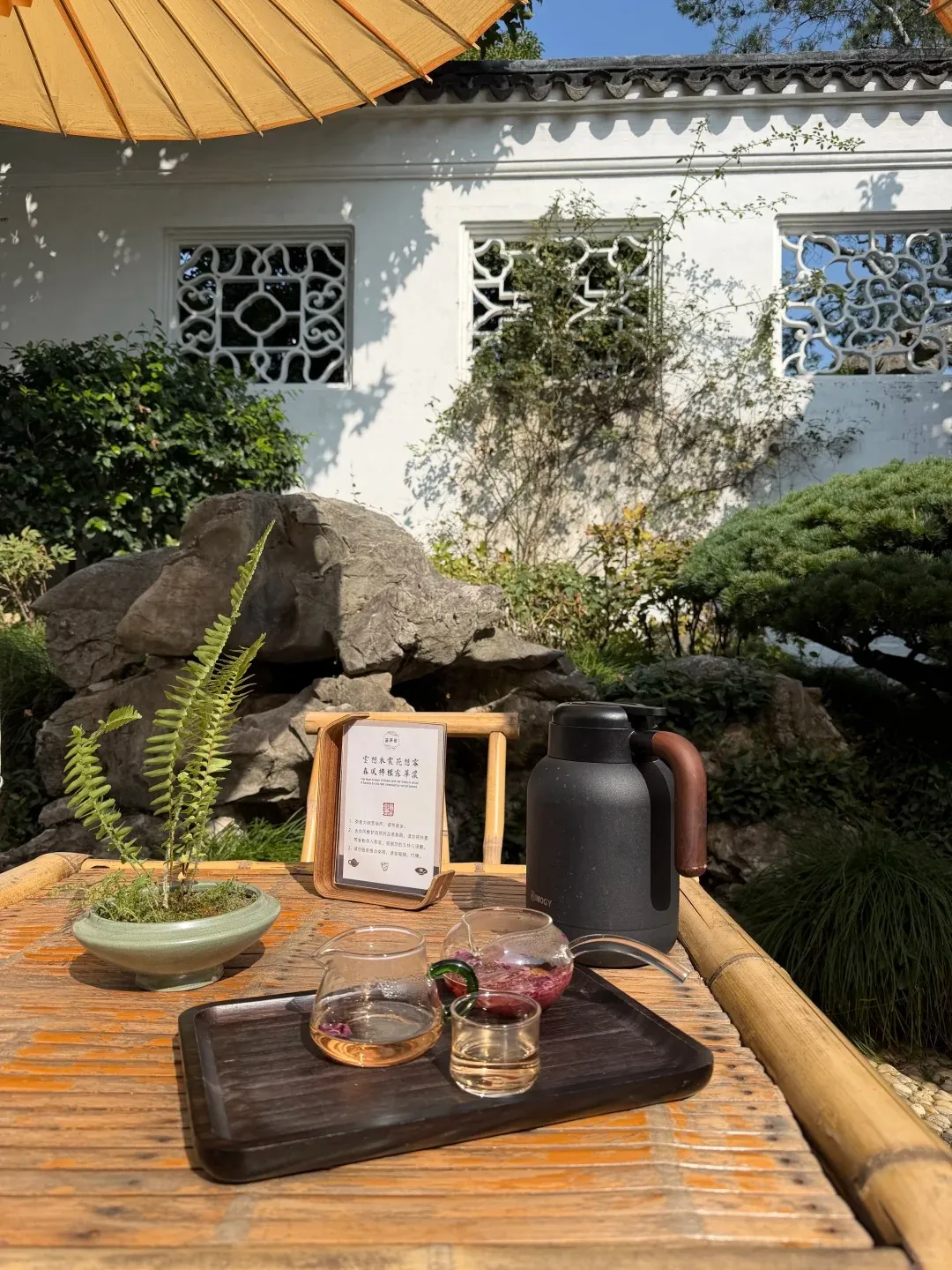 An outdoor bamboo table with a dark wooden tray holding a glass teapot of rose tea, a glass pitcher of clear tea, and a small glass cup. A potted fern and a black insulated bottle are also on the table. In the background, large rocks, green foliage, and a white traditional wall with two decorative carved windows are visible under a bright sky.